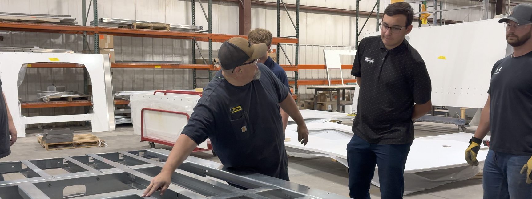 A warehouse or fabrication area. A man in a brown hat wearing a carhart tee with embroidered Marion logo points to a metal frame. Three young men observe with interest. The young man in the foreground wears glasses and a black polo embroidered with a white Marion logo.