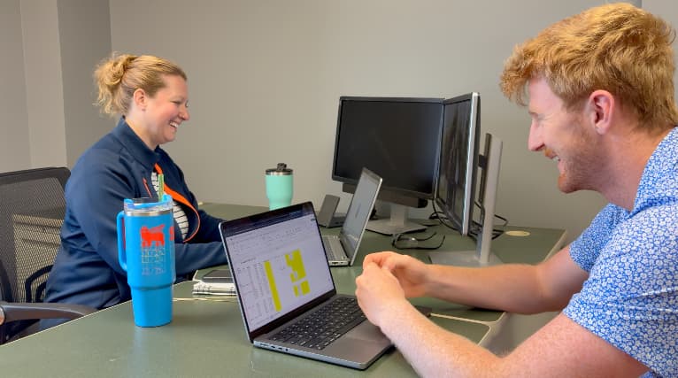 A man and woman meeting at an office desk. They are seated across from each other in front of separate laptops with the screens open. A large blue mug is on the desk in the foreground.