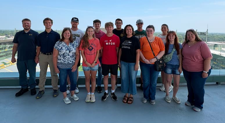 14 people standing posed for a photo at a high vantage point on the south end of Lambeau Field with the stadium, scoreboard, and neighborhood buildings and trees in the background.