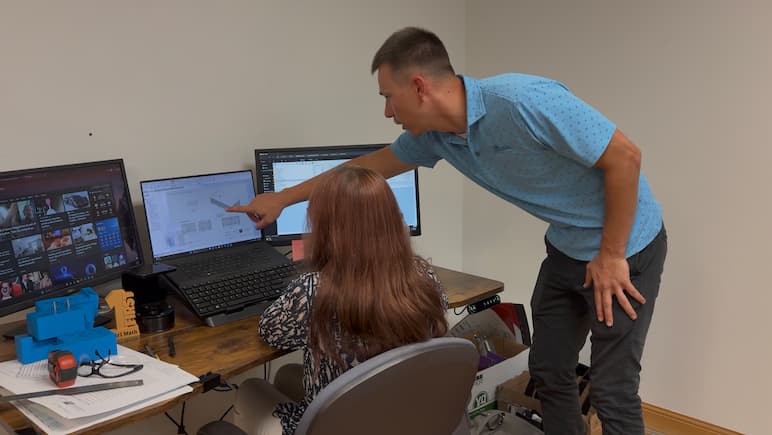 Office workers collaborating on a digital project. From this camera angle we see the back of a woman's head and chair. The woman is seated at an office desk in front of a laptop and 2 external monitors. A man stands to the right in a blue polo shirt pointing to the laptop screen.